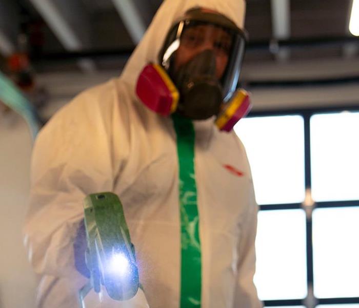 A man in a white suit, gloves, and mask spraying disinfectant from a green fogger.