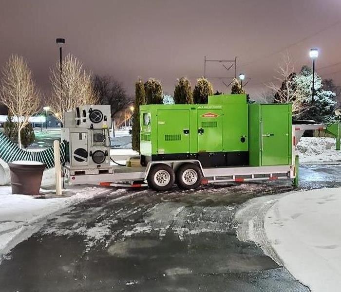 A green semi trailer with a large green machine and generator sitting on it parked in front of a hotel.